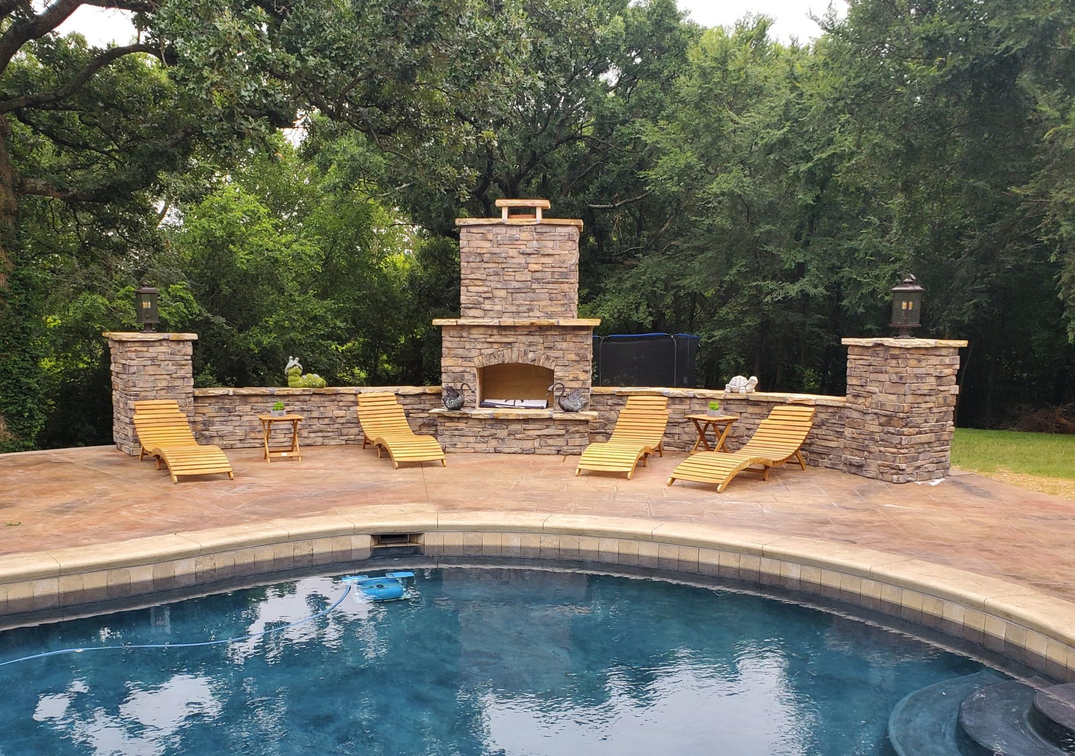 Stone patio with loungers and fireplace next to an inground swimming pool.