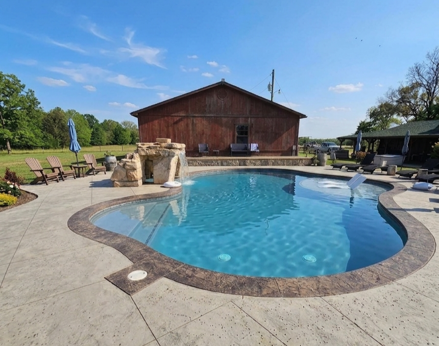 Curved inground swimming pool with stone waterfall near a rustic wooden building.