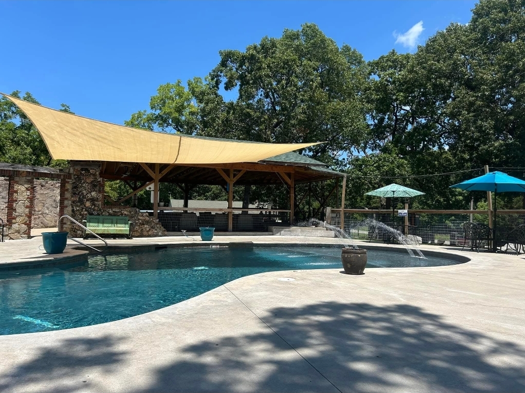 Outdoor swimming pool with water fountains and shaded seating area.